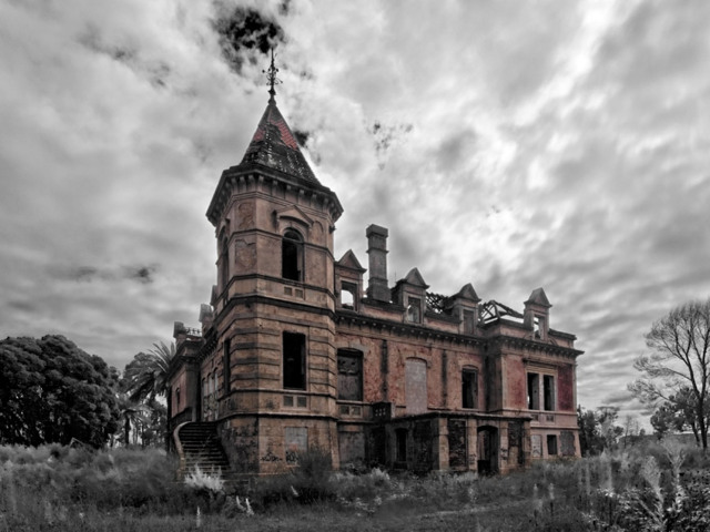 The image shows a spooky place in Portugal. This is Palácio Marques Gomes in Gaia. The image shows the palace in color and the surroundings in black and white. The weather is very cloudy.  Around the palace there is only nature, trees and low greens. The palace is in red brick and looks scary and abandoned. There are window openings without glass on it. The palace has a tower in the left front corner.