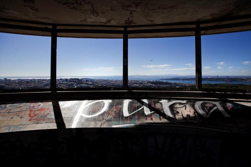 The image shows a spooky place in Lisboa. There is a panoramic view over Lisbon and the Tagus River. We look into it through four glass-less window openings and a tiled illustration of Lisbon's most iconic buildings is below the openings. The weather is sunny and clear sky.