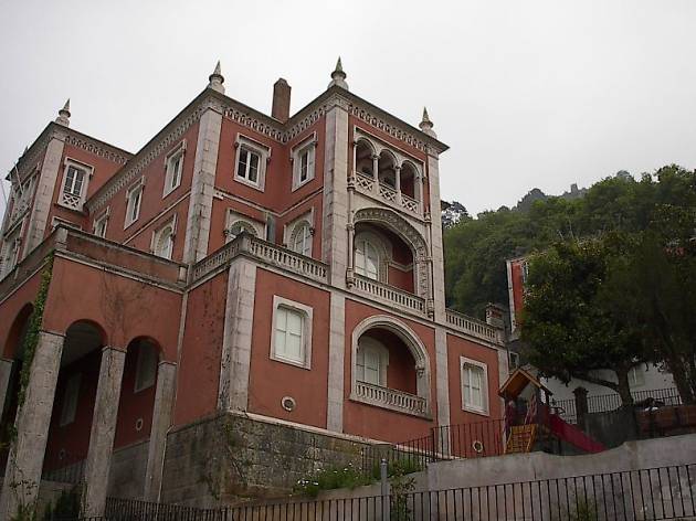 The image shows a spooky place in Portugal. There is a palace seen from down up, from the street. The palace is in a dark pink-reddish color, with corners in limestone. The weather is cloudy.