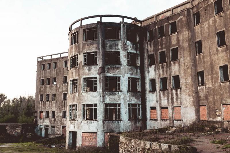 The image shows a spooky place in Portugal. it is an abandoned sanatorium in Valongo, in the north of Portugal.  The place looks abandoned, with dirty walls, broken glass and empty window and door openings. Surrounding it there is nature. The weather is cloudy with some sunset lighting.